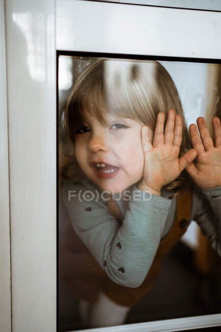 Мила дівчинка в повсякденному одязі дивиться на камеру і посміхається, стоячи за вікном вдома і торкаючись скла — стокове фото