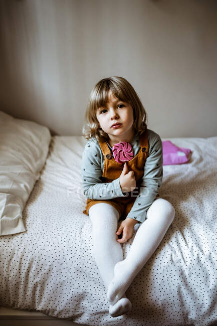 Cute little girl in casual clothes looking at camera and sucking lollipop while sitting on comfortable bed near toy at home — Stock Photo