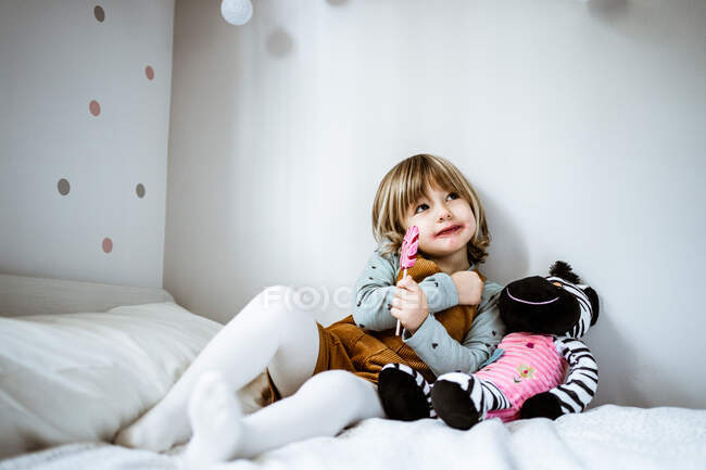 Linda niña en ropa casual mirando a la cámara y chupando piruleta mientras está sentada en una cama cómoda cerca de un juguete en casa - foto de stock