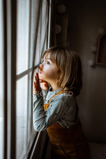 Adorable petite fille en vêtements décontractés regardant par la fenêtre tout en se reposant dans une chambre confortable à la maison — Photo de stock