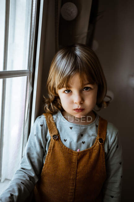 Adorable niña en ropa casual mirando por la ventana mientras descansa en la acogedora habitación en casa - foto de stock