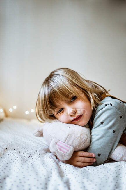 Возбужденная маленькая девочка, обнимающая плюшевую игрушку и смеющаяся, лежа на мягкой кровати возле волшебных огней дома — стоковое фото