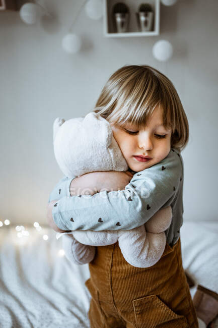 Emocionado menina abraçando brinquedo de pelúcia e rindo enquanto deitado em cama macia perto de luzes de fadas em casa — Fotografia de Stock