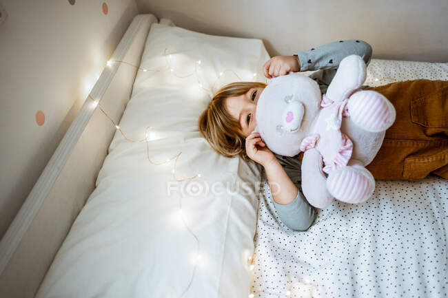 Aufgeregtes kleines Mädchen umarmt Plüschtier und lacht, während es auf weichem Bett in der Nähe von Lichterketten zu Hause liegt — Stockfoto
