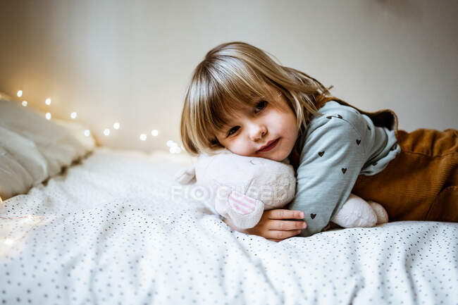 Aufgeregtes kleines Mädchen umarmt Plüschtier und lacht, während es auf weichem Bett in der Nähe von Lichterketten zu Hause liegt — Stockfoto