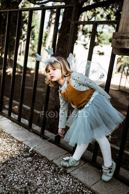 Adorable little girl in fairy costume sitting on shabby wooden bench and looking at camera while spending time in yard — Stock Photo