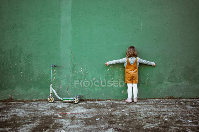 Back view of anonymous little girl with outstretched arms standing near weathered green wall and kick scooter and counting while playing hide and seek — Stock Photo