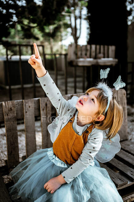Чарівна маленька дівчинка в казковому костюмі, сидячи на похмурій дерев'яній лавці і дивлячись на камеру, проводячи час у дворі — стокове фото