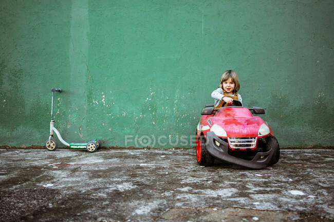 Nettes kleines Mädchen in lässiger Kleidung schaut in die Kamera und fährt rotes Elektrofahrzeug mit kaputter Stoßstange gegen schäbige grüne Wand in der Nähe von Tretroller — Stockfoto