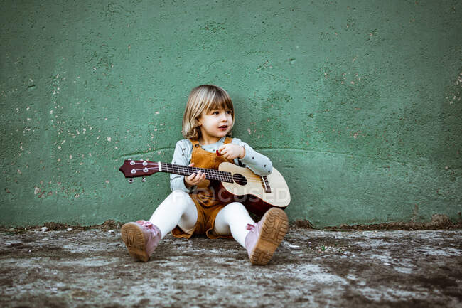 Menina com ukulele sentado em solo áspero perto de chutar scooter contra parede verde resistido na rua — Fotografia de Stock