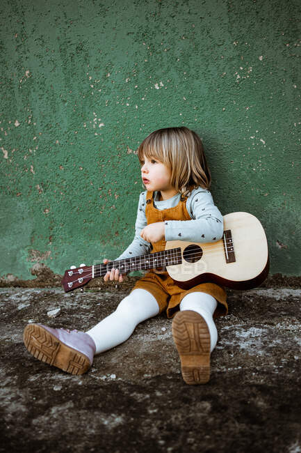 Petite fille avec ukulélé assis sur un terrain accidenté près de trottinette de coup de pied contre le mur vert altéré sur la rue — Photo de stock
