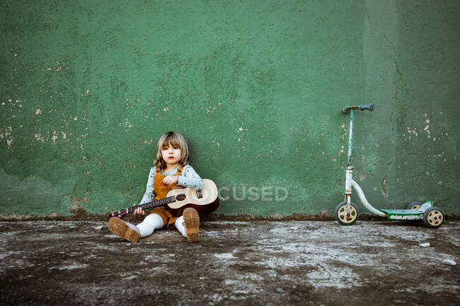 Bambina con ukulele seduta su terreno accidentato vicino al calcio scooter contro il muro verde intemperie sulla strada — Foto stock