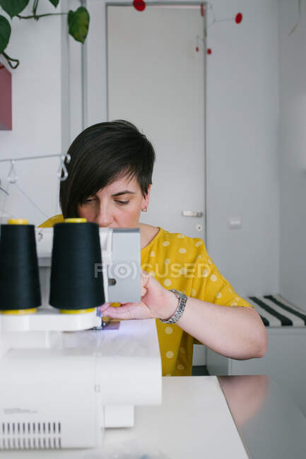 Сфокусована на брюнетці доросла жінка посміхається і використовує швейну машину для виготовлення одягу під час роботи в домашній майстерні. — стокове фото