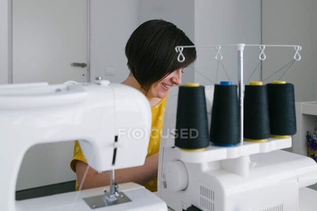 Glückliche brünette erwachsene Frau lächelt und verwendet Nähmaschine, um Kleidungsstücke zu machen, während sie in der heimischen Werkstatt arbeitet — Stockfoto
