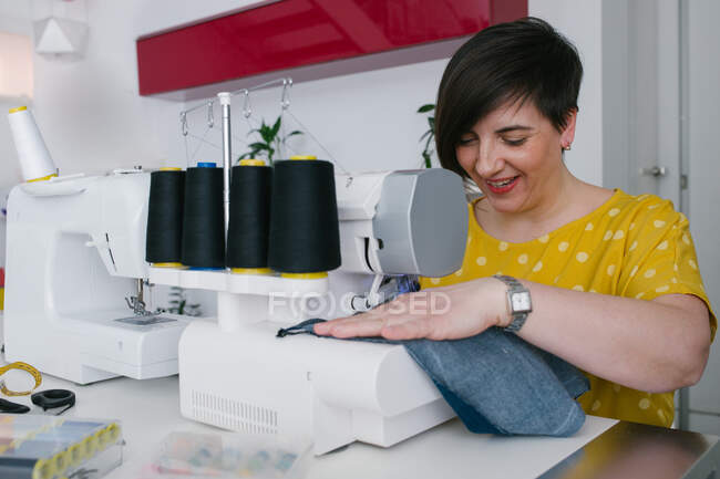 Feliz morena mujer adulta sonriendo y utilizando la máquina de coser para hacer prendas de mezclilla mientras trabaja en el taller en casa - foto de stock