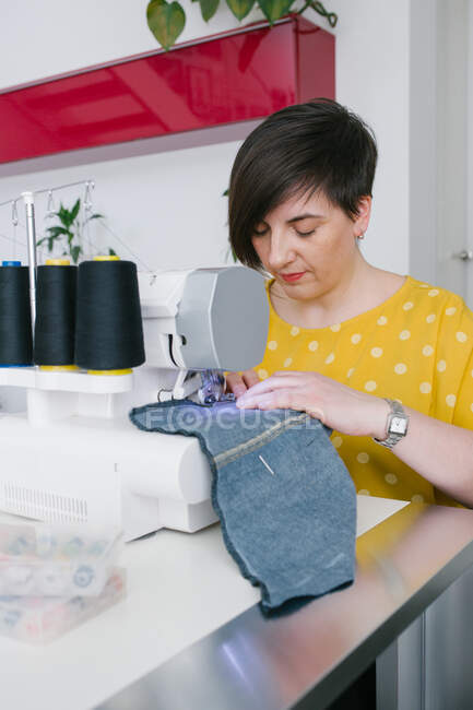 Konzentrierte brünette erwachsene Frau lächelt und verwendet Nähmaschine, um Jeans-Kleidungsstück zu machen, während sie in der heimischen Werkstatt arbeitet — Stockfoto