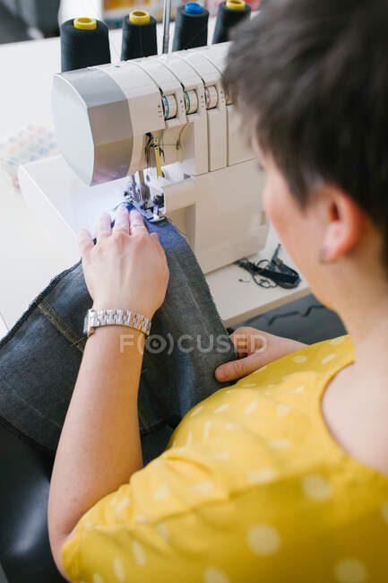 Dall'alto ritagliato donna adulta bruna irriconoscibile utilizzando la macchina da cucire per fare indumento in denim mentre si lavora in officina a casa — Foto stock