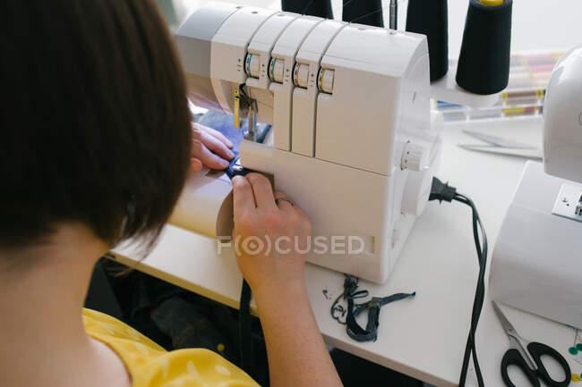 Desde arriba recortado irreconocible morena mujer adulta utilizando la máquina de coser para hacer la prenda de mezclilla mientras trabaja en el taller en casa - foto de stock