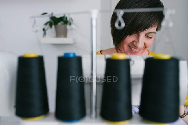 Щаслива брюнетка доросла жінка посміхається і використовує швейну машину для виготовлення одягу під час роботи в домашній майстерні — стокове фото