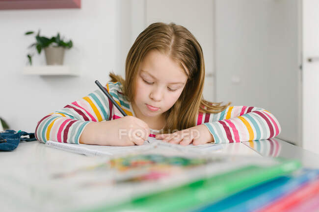 Focused girl in striped t shirt writing in notebook while sitting at table and doing homework at home — Stock Photo