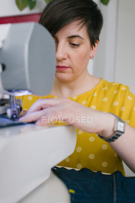 Blurred brunette adult woman using sewing machine to make denim garment while working in home workshop — Stock Photo