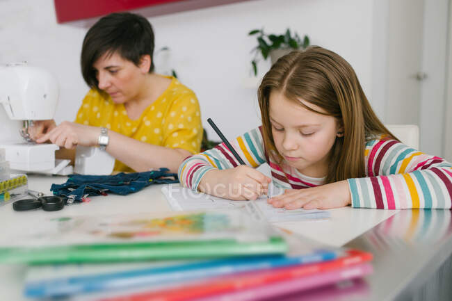 Focused girl doing homework assignment while sitting near adult woman sewing garment at home — Stock Photo