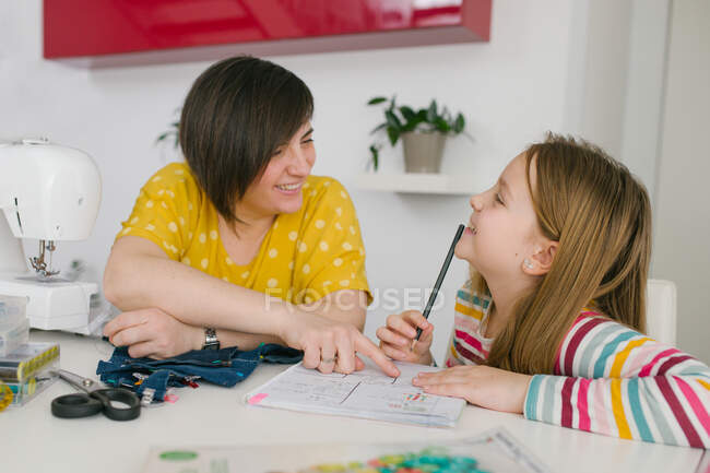 Happy adult woman smiling and helping girl with homework assignment while sewing garment at home — Stock Photo