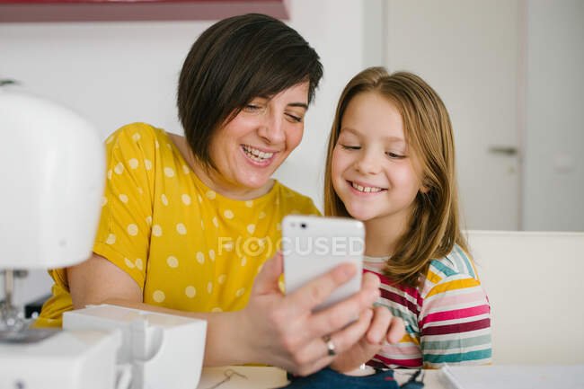 Femme adulte joyeuse souriant et prenant selfie avec la fille tout en étant assis à la table et travaillant dans l'atelier de couture à la maison — Photo de stock