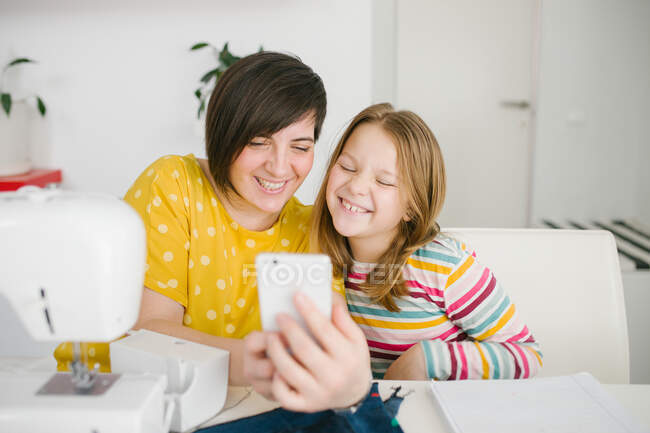 Mujer adulta alegre sonriendo y tomando selfie con la chica mientras está sentado en la mesa y trabajando en taller de costura en casa - foto de stock