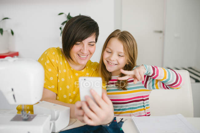Mujer adulta alegre sonriendo y tomando selfie con la chica mientras está sentado en la mesa y trabajando en taller de costura en casa - foto de stock
