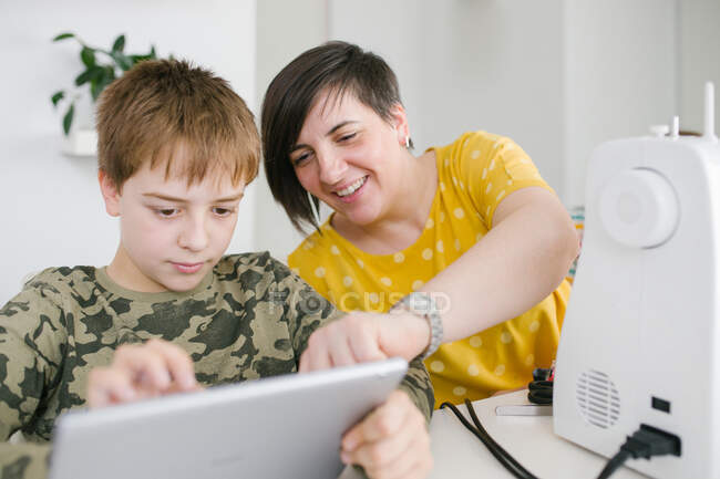 Взрослая женщина помогает маленькому мальчику просматривать современные планшеты вместе, сидя за столом дома — стоковое фото
