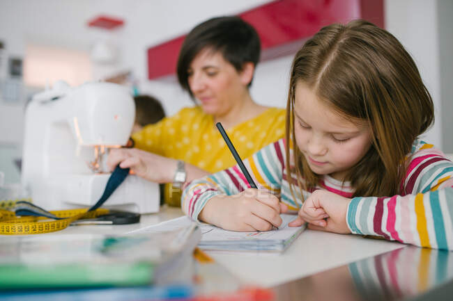 Focused girl doing homework assignment while sitting near adult woman sewing garment at home — Stock Photo