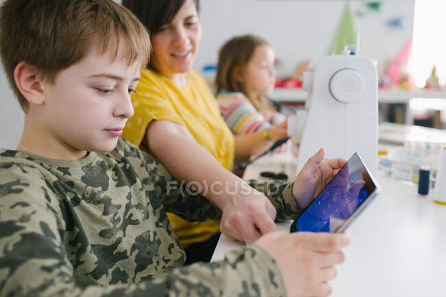 Mujer adulta ayudando a niño pequeño navegar tableta moderna juntos mientras están sentados en la mesa en casa - foto de stock