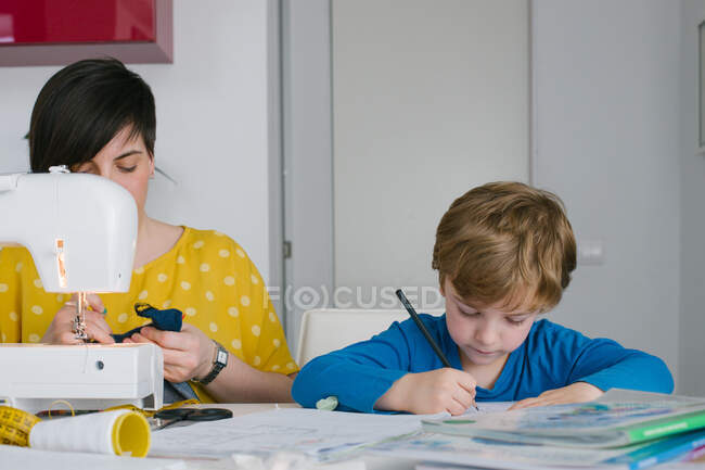 Сосредоточенный мальчик делает домашнее задание, сидя рядом со взрослой женщиной, шьет одежду дома — стоковое фото