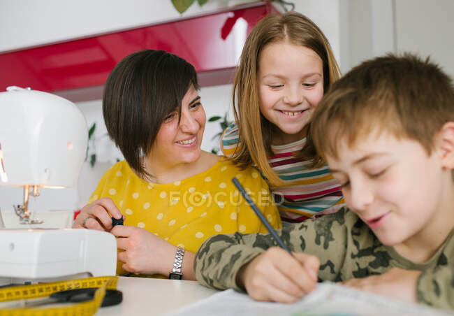 Heureuse femme adulte et fille souriant et regardant le garçon faire ses devoirs tout en passant du temps dans le studio de couture à la maison ensemble — Photo de stock