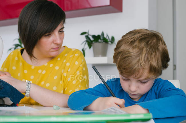 Focused ragazzo fare compiti a casa assegnazione mentre seduto vicino a donna adulta cucire indumento a casa — Foto stock