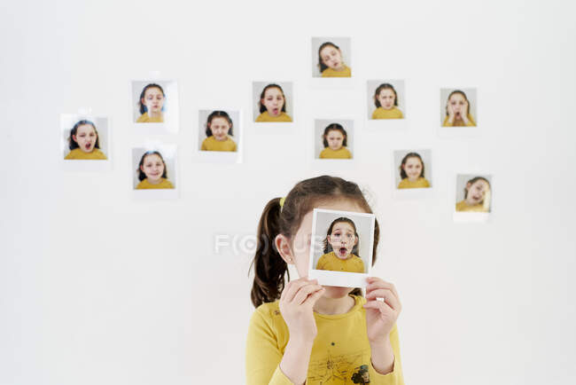 Cute little girl in yellow dress hiding face behind own picture while standing against wall with photos demonstrating various emotions — Stock Photo
