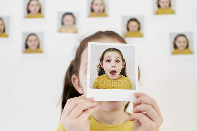 Nettes kleines Mädchen in gelbem Kleid versteckt Gesicht hinter eigenem Bild, während es an der Wand steht und Fotos zeigt, die verschiedene Emotionen zeigen — Stockfoto