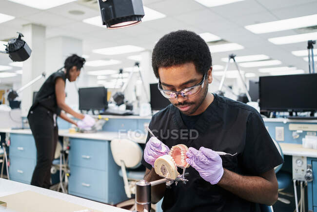 Chico afroamericano en guantes de látex usando espejo bucal y sonda para comprobar dientes falsos mientras trabaja en un laboratorio moderno - foto de stock