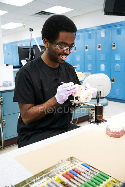 Chico afroamericano en guantes de látex usando espejo bucal y sonda para comprobar dientes falsos mientras trabaja en un laboratorio moderno - foto de stock