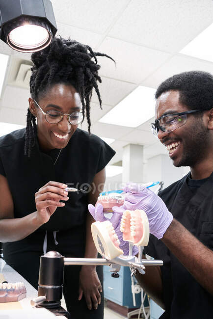 Низький кут щасливої афро-американської жінки з дредами і бородатий чоловік посміхається і чистить фальшиві зуби під час роботи в лабораторії — стокове фото