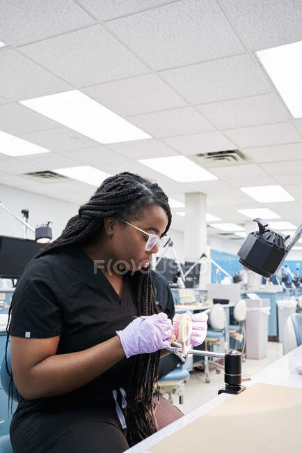 Délicieuse femme afro-américaine avec des tresses utilisant un miroir buccal et une sonde pour montrer à ses collègues de fausses dents pendant le travail en laboratoire — Photo de stock
