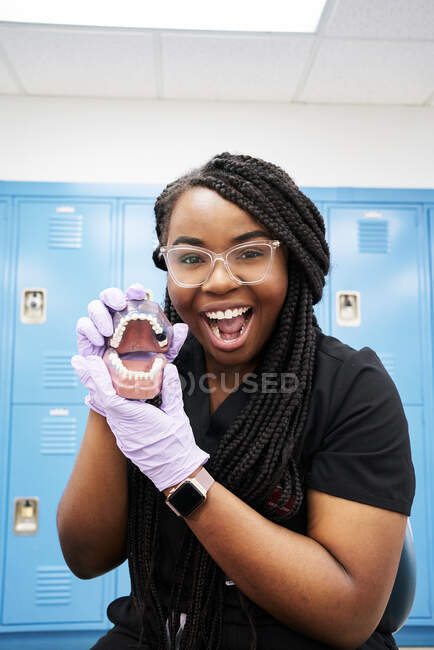 Щасливий чорний стоматолог з косами дивиться на камеру і показує фальшиві зуби під час роботи в сучасній лабораторії — стокове фото