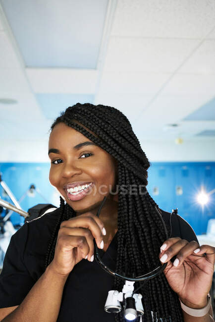 Mujer negra feliz con trenzas sonriendo y mirando a la cámara antes de ponerse gafas binoculares durante el trabajo en el laboratorio contemporáneo - foto de stock