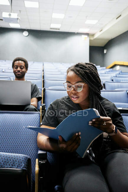 Afroamerikanerin mit Brille liest Notizen, während sie vor dem Vortrag im modernen Hörsaal neben einem schwarzen Mann mit Laptop sitzt — Stockfoto