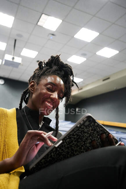 Desde abajo mujer negra con rastas sonriendo y usando la tableta mientras se sienta en la sala de conferencias iluminada durante la lección - foto de stock