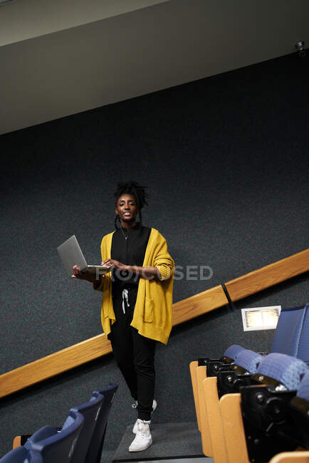 Full body African American female in casual clothes looking at camera and browsing laptop while standing amidst rows of seats in modern lecture hall — Stock Photo