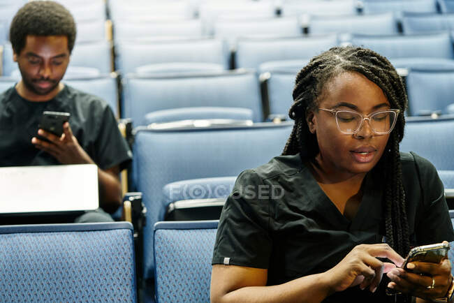 Mulher negra com tranças e homem afro-americano sentado no auditório e navegando smartphones durante a aula de auditório — Fotografia de Stock