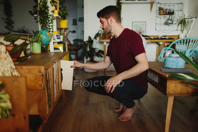 Бічний вид на босоногого чоловіка, який сидить на переслідуваннях і збирає вініловий диск з полиці в затишній кімнаті вдома. — стокове фото