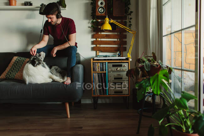 Cara em fones de ouvido ouvindo música na plataforma giratória e brincando com Collie enquanto sentado no sofá em casa — Fotografia de Stock